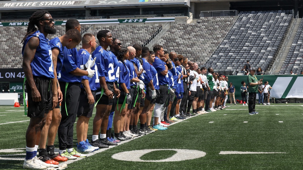A large group of football players in blue jerseys are lined up on the field at MetLife Stadium, home of the New York Jets. The players appear to be standing at attention, likely during a pre-game ceremony such as the national anthem. The stadium seats are largely empty, suggesting this may be a practice, scrimmage, or pre-season event.