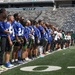 A large group of football players in blue jerseys are lined up on the field at MetLife Stadium, home of the New York Jets. The players appear to be standing at attention, likely during a pre-game ceremony such as the national anthem. The stadium seats are largely empty, suggesting this may be a practice, scrimmage, or pre-season event.
