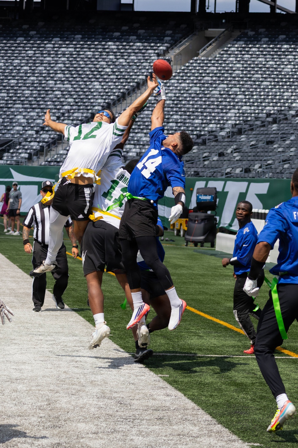 A flag football game is being played on the field of MetLife Stadium, home of the New York Jets, as indicated by the visible 'JETS' signage on the wall. Multiple players are jumping to contest a football near the sideline, with players in white/green jerseys competing against players in blue jerseys. The stadium seats are largely empty, suggesting this is a non-game-day event or youth/amateur competition.