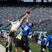 A flag football game is being played on the field of MetLife Stadium, home of the New York Jets, as indicated by the visible 'JETS' signage on the wall. Multiple players are jumping to contest a football near the sideline, with players in white/green jerseys competing against players in blue jerseys. The stadium seats are largely empty, suggesting this is a non-game-day event or youth/amateur competition.