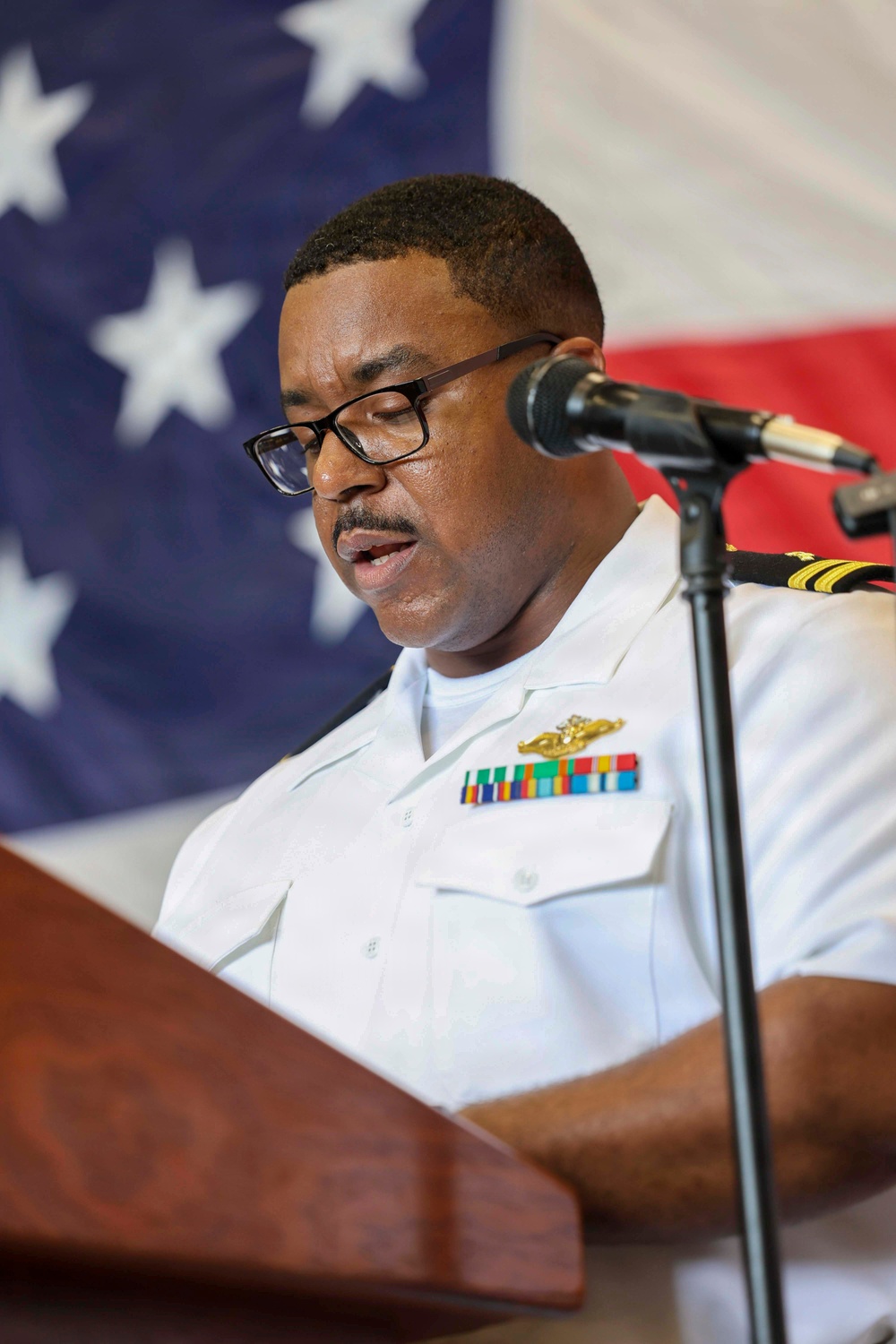 A U.S. Navy officer in white service uniform is speaking at a wooden podium with a microphone. He is wearing glasses and has multiple service ribbons and a warfare qualification badge on his chest. An American flag is displayed prominently in the background.