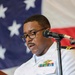 A U.S. Navy officer in white service uniform is speaking at a wooden podium with a microphone. He is wearing glasses and has multiple service ribbons and a warfare qualification badge on his chest. An American flag is displayed prominently in the background.