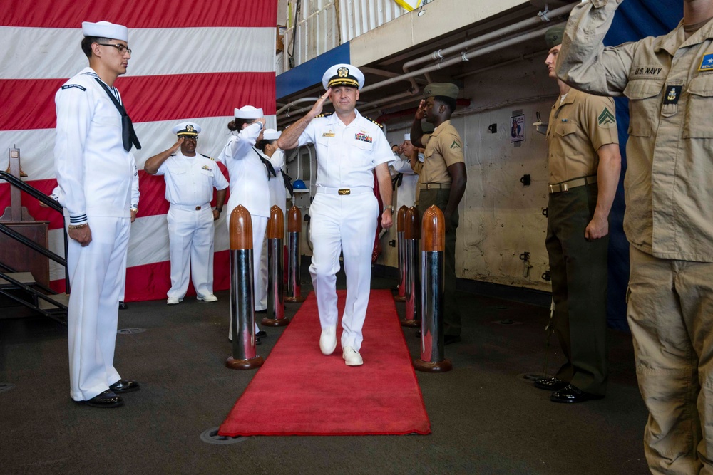 A U.S. Navy officer in dress white uniform walks a red carpet during a formal military ceremony, likely a change of command or sideboy honors ritual. The officer is rendering a salute while passing through a sideboy formation flanked by decorative shell casings on wooden bases. Personnel in various uniforms, including Navy whites and tan/khaki working uniforms, are present and saluting.