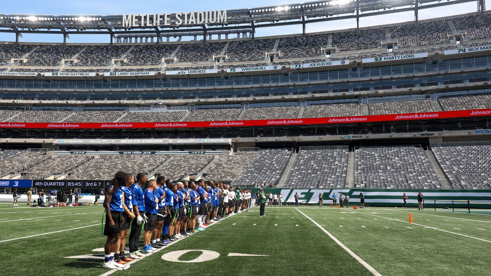 A group of youth football players in blue uniforms are lined up on the field at MetLife Stadium, home of the New York Jets and Giants. The stadium is largely empty, suggesting this is a special event or youth program activity rather than a professional game. The scoreboard displays '4th Quarter 85:45 3rd &amp; 7' which appears to be part of a youth game or event.