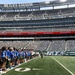 A group of youth football players in blue uniforms are lined up on the field at MetLife Stadium, home of the New York Jets and Giants. The stadium is largely empty, suggesting this is a special event or youth program activity rather than a professional game. The scoreboard displays '4th Quarter 85:45 3rd &amp; 7' which appears to be part of a youth game or event.