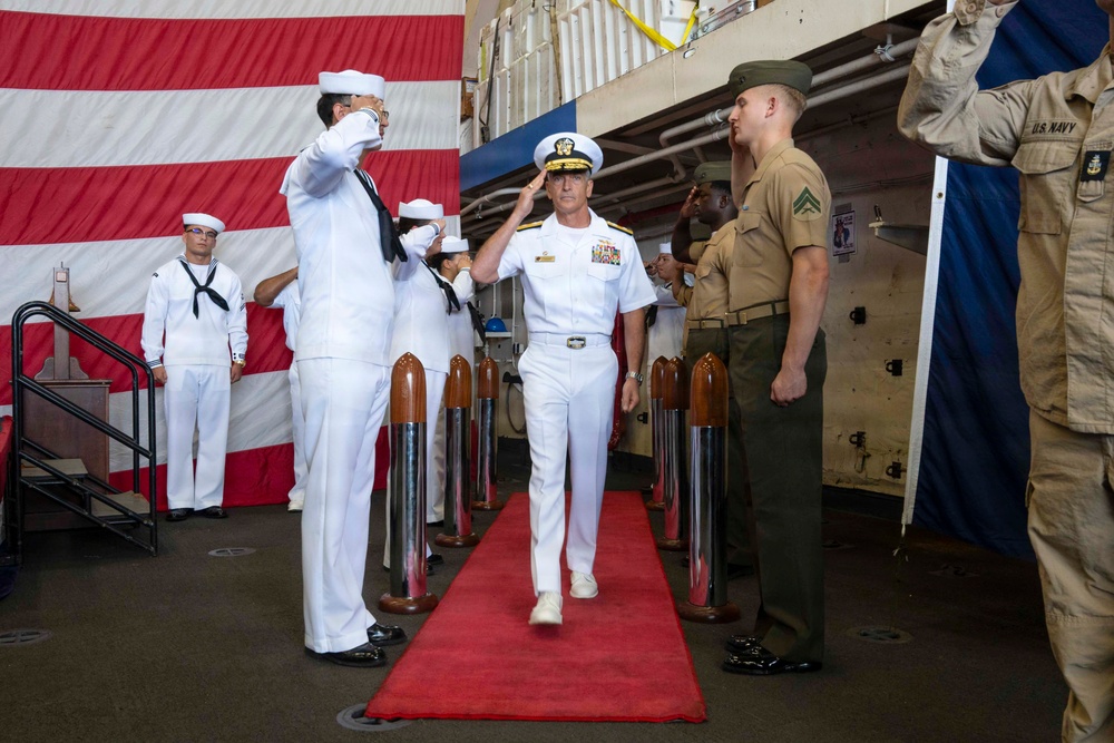 A U.S. Navy admiral in dress white uniform walks along a red carpet through a sideboy honors ceremony, being saluted by a row of Navy sailors in white uniforms and Marine Corps personnel in service uniforms. The ceremony appears to be taking place in a ship's hangar bay, decorated with a large American flag in the background. This is consistent with a formal military change of command or arrival ceremony aboard a U.S. Navy vessel.