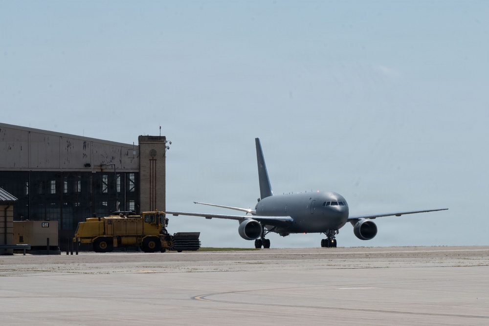 A large gray military aircraft, consistent with a KC-46 Pegasus or similar tanker/transport, is taxiing on an airfield apron near a large industrial hangar. A yellow Caterpillar (CAT) ground support vehicle is parked adjacent to the hangar building. The scene appears to be at a military air base under overcast, hazy sky conditions.