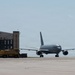 A large gray military aircraft, consistent with a KC-46 Pegasus or similar tanker/transport, is taxiing on an airfield apron near a large industrial hangar. A yellow Caterpillar (CAT) ground support vehicle is parked adjacent to the hangar building. The scene appears to be at a military air base under overcast, hazy sky conditions.