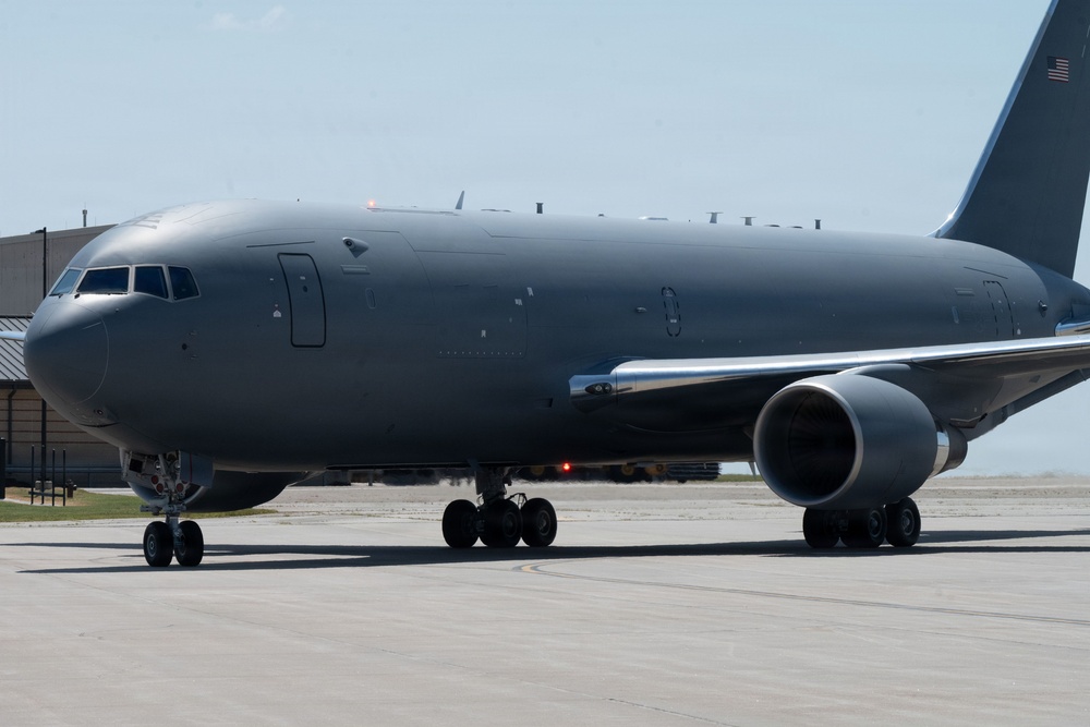 A large military aircraft painted in matte gray is taxiing on an airfield tarmac. The aircraft appears to be a Boeing KC-46 Pegasus or similar military tanker/transport variant, identifiable by its wide-body fuselage and military gray livery. An American flag is visible on the tail section, confirming U.S. military ownership.