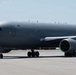 A large military aircraft painted in matte gray is taxiing on an airfield tarmac. The aircraft appears to be a Boeing KC-46 Pegasus or similar military tanker/transport variant, identifiable by its wide-body fuselage and military gray livery. An American flag is visible on the tail section, confirming U.S. military ownership.