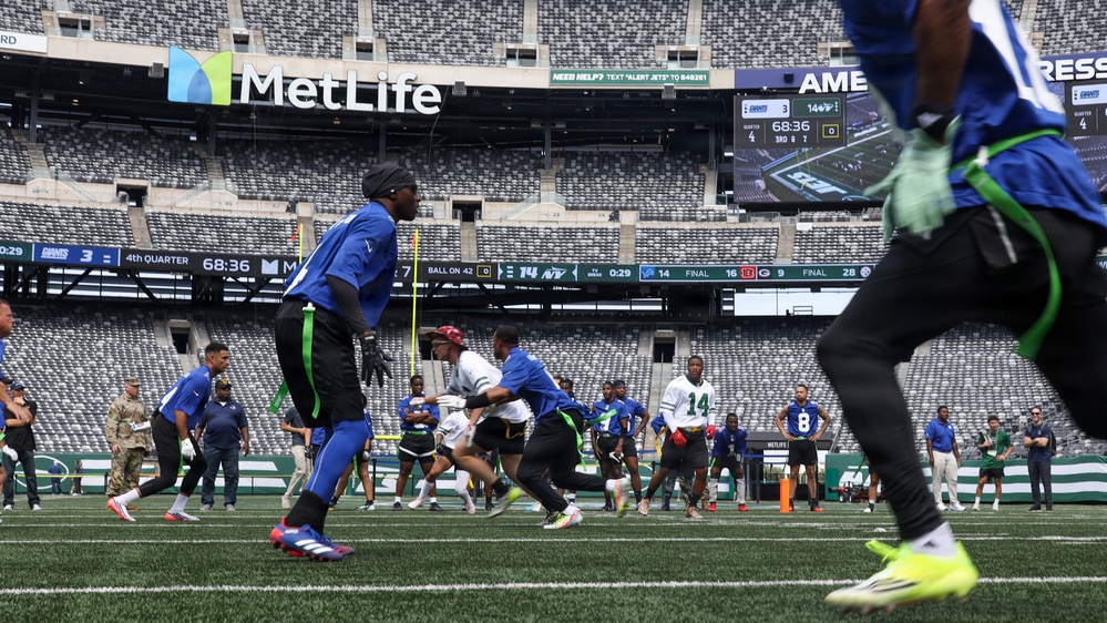 A flag football game is being played on the field at MetLife Stadium, home of the New York Giants and Jets. Players in blue and white/green uniforms are actively competing on the turf while spectators and staff watch from the sidelines. The stadium scoreboard shows a 4th quarter game situation with 0:29 remaining.