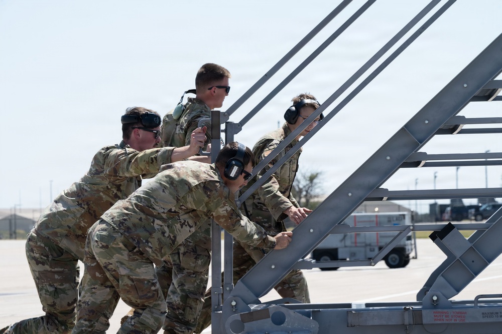 A group of military personnel in camouflage uniforms are collectively pushing or maneuvering a metal aircraft boarding staircase on what appears to be a military airfield flight line. The personnel are wearing hearing protection and some have eye protection, indicating proximity to aircraft operations. The setting appears to be a sunny day at an active airbase with ground support equipment visible in the background.