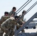 A group of military personnel in camouflage uniforms are collectively pushing or maneuvering a metal aircraft boarding staircase on what appears to be a military airfield flight line. The personnel are wearing hearing protection and some have eye protection, indicating proximity to aircraft operations. The setting appears to be a sunny day at an active airbase with ground support equipment visible in the background.
