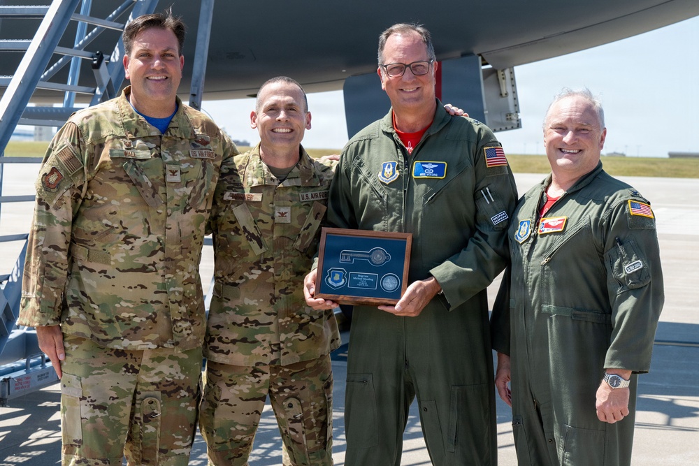 Four U.S. Air Force personnel are posing for a photo on an airfield tarmac in front of what appears to be a large military aircraft. The central figure is holding a ceremonial plaque featuring a key design, military unit patches, and an inscription, suggesting a formal recognition or retirement ceremony. Two individuals are wearing OCP (Operational Camouflage Pattern) uniforms while two are wearing green flight suits.