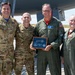 Four U.S. Air Force personnel are posing for a photo on an airfield tarmac in front of what appears to be a large military aircraft. The central figure is holding a ceremonial plaque featuring a key design, military unit patches, and an inscription, suggesting a formal recognition or retirement ceremony. Two individuals are wearing OCP (Operational Camouflage Pattern) uniforms while two are wearing green flight suits.