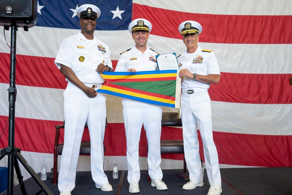 Three U.S. Navy personnel in white dress uniforms are posing together while holding a multi-colored streamer/pennant flag during what appears to be a formal military ceremony. The individual in the center is also holding what appears to be an official document or citation. A large American flag serves as the backdrop for the ceremony.