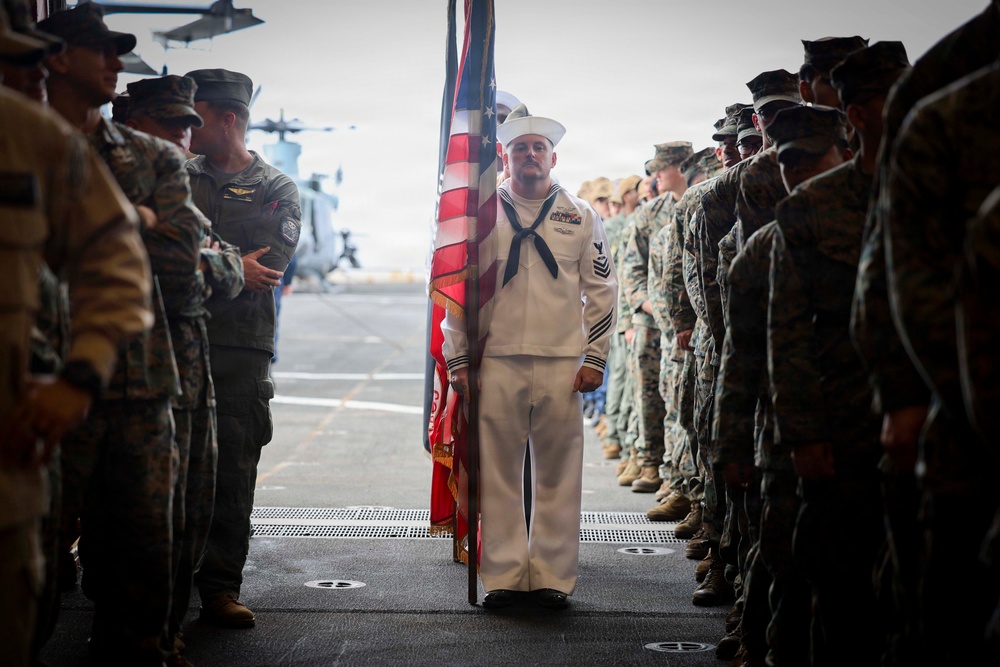A U.S. Navy sailor in dress white uniform stands at the center holding an American flag on what appears to be the flight deck of a naval vessel. He is flanked on both sides by rows of U.S. Marines in camouflage combat uniforms standing at attention. A military helicopter is visible in the background, suggesting this is a formal ceremony aboard a ship.