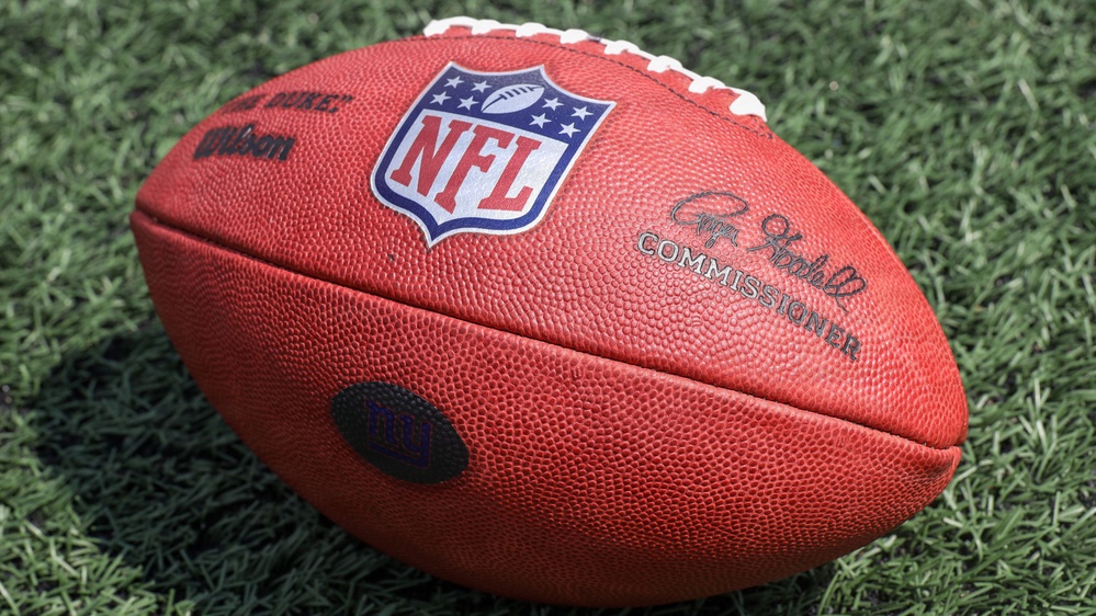 A close-up photograph of an official NFL Wilson Duke football resting on green artificial turf. The ball displays the NFL shield logo, Wilson branding, and the Commissioner's signature. A team logo stamp is partially visible on the side of the ball.