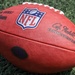 A close-up photograph of an official NFL Wilson Duke football resting on green artificial turf. The ball displays the NFL shield logo, Wilson branding, and the Commissioner's signature. A team logo stamp is partially visible on the side of the ball.