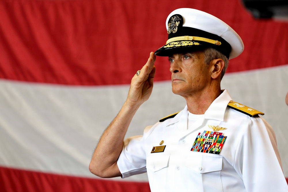 A senior U.S. Navy flag officer in white dress uniform is rendering a formal salute against a backdrop of red and white bunting or drapery. The officer displays multiple rows of service ribbons, gold aviator wings, and shoulder boards indicating high flag rank. The setting appears to be a formal military ceremony.