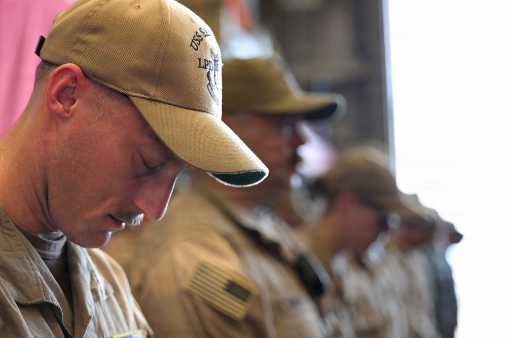 A group of military personnel in tan/khaki uniforms are standing in a line with their heads bowed, suggesting a moment of silence, prayer, or solemn ceremony. The foreground subject wears a baseball cap with 'USS' and 'LPD' visible, indicating U.S. Navy affiliation. An American flag patch is visible on the sleeve of the foreground individual.