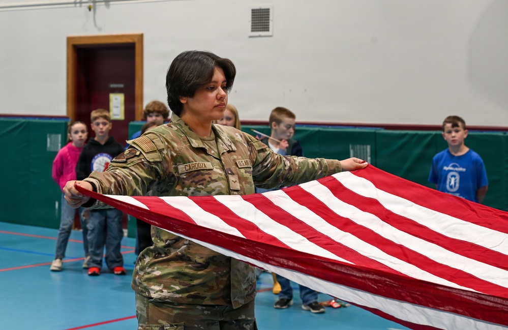 A U.S. Army soldier named Cardoza, wearing OCP (Operational Camouflage Pattern) uniform, is demonstrating American flag folding to a group of elementary school children in a gymnasium. The soldier holds the flag stretched out horizontally while children observe attentively in the background. This appears to be an educational outreach or Veterans Day-type school visit.
