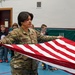 A U.S. Army soldier named Cardoza, wearing OCP (Operational Camouflage Pattern) uniform, is demonstrating American flag folding to a group of elementary school children in a gymnasium. The soldier holds the flag stretched out horizontally while children observe attentively in the background. This appears to be an educational outreach or Veterans Day-type school visit.