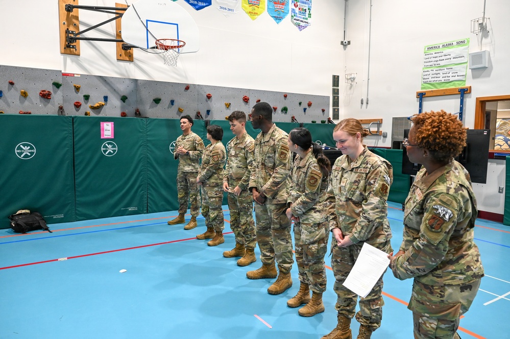 A group of military personnel in OCP (Operational Camouflage Pattern) uniforms are assembled in a school gymnasium, appearing to be in a formation or briefing. A senior service member on the right is holding papers and appears to be addressing or reviewing the group. The setting is identifiable as a school gym in Alaska based on visible signage including an 'America Alaska Song' poster.