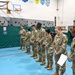 A group of military personnel in OCP (Operational Camouflage Pattern) uniforms are assembled in a school gymnasium, appearing to be in a formation or briefing. A senior service member on the right is holding papers and appears to be addressing or reviewing the group. The setting is identifiable as a school gym in Alaska based on visible signage including an 'America Alaska Song' poster.