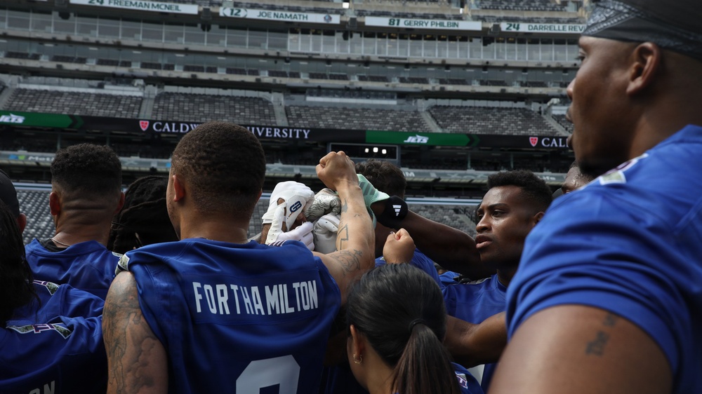 A group of football players in blue jerseys are gathered in a team huddle or celebration on a stadium field. The stadium backdrop shows retired jersey numbers including Joe Namath (#12) and other New York Jets legends, along with a 'Caldwell University' banner visible on the scoreboard. The players have their fists raised in a show of team unity.