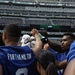 A group of football players in blue jerseys are gathered in a team huddle or celebration on a stadium field. The stadium backdrop shows retired jersey numbers including Joe Namath (#12) and other New York Jets legends, along with a 'Caldwell University' banner visible on the scoreboard. The players have their fists raised in a show of team unity.