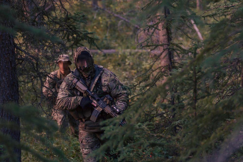 Two armed individuals in full camouflage military-style gear are moving through a dense coniferous forest. The foreground figure is heavily equipped with a face covering, hood, tactical vest, and an assault-style rifle. The background figure wears a boonie hat and similar camouflage uniform.
