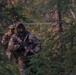 Two armed individuals in full camouflage military-style gear are moving through a dense coniferous forest. The foreground figure is heavily equipped with a face covering, hood, tactical vest, and an assault-style rifle. The background figure wears a boonie hat and similar camouflage uniform.