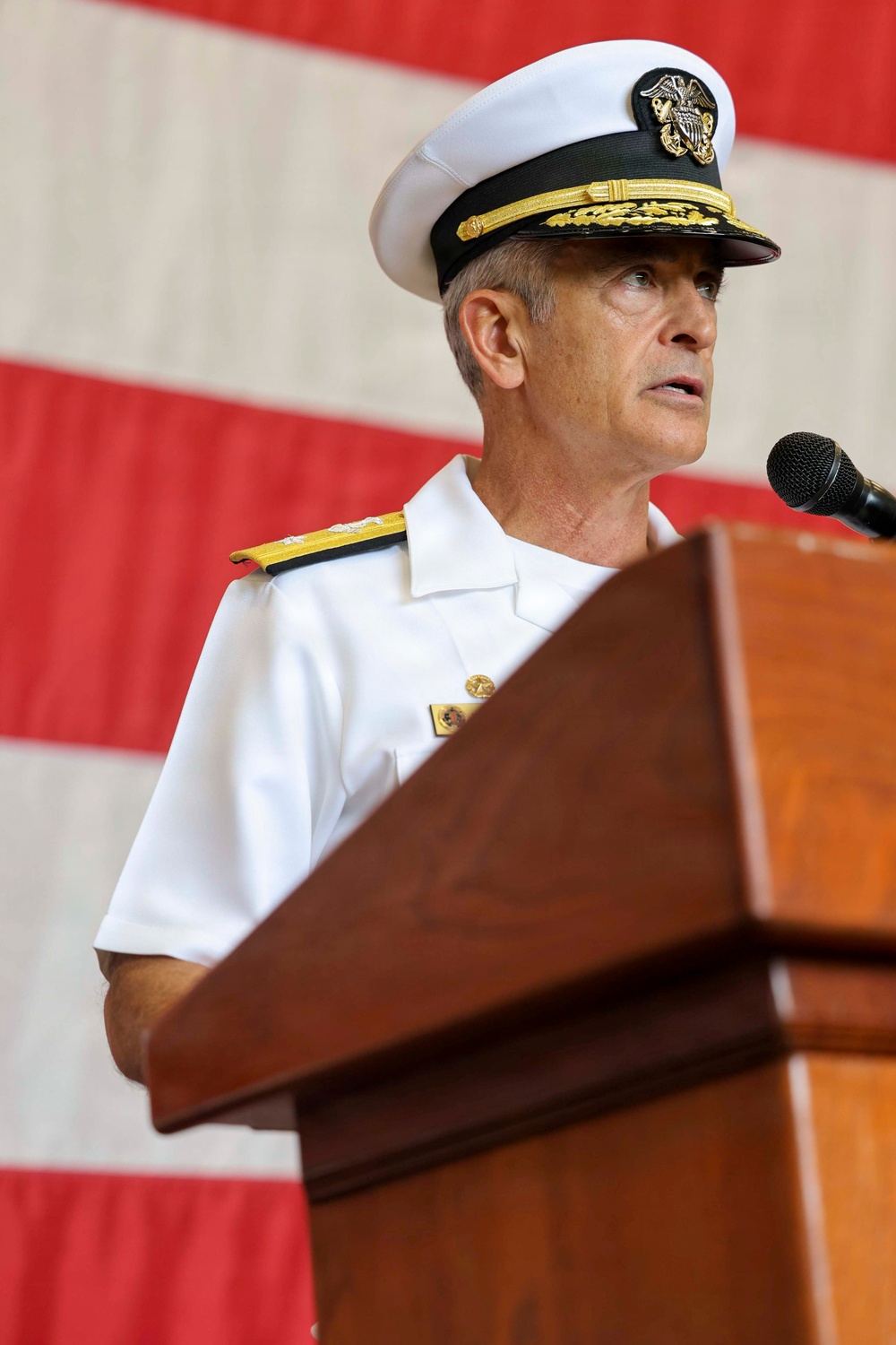 A senior U.S. Navy officer in white service dress uniform stands at a wooden podium delivering a speech. The background features a large American flag with visible red and white stripes. The setting appears to be a formal military ceremony or official event.