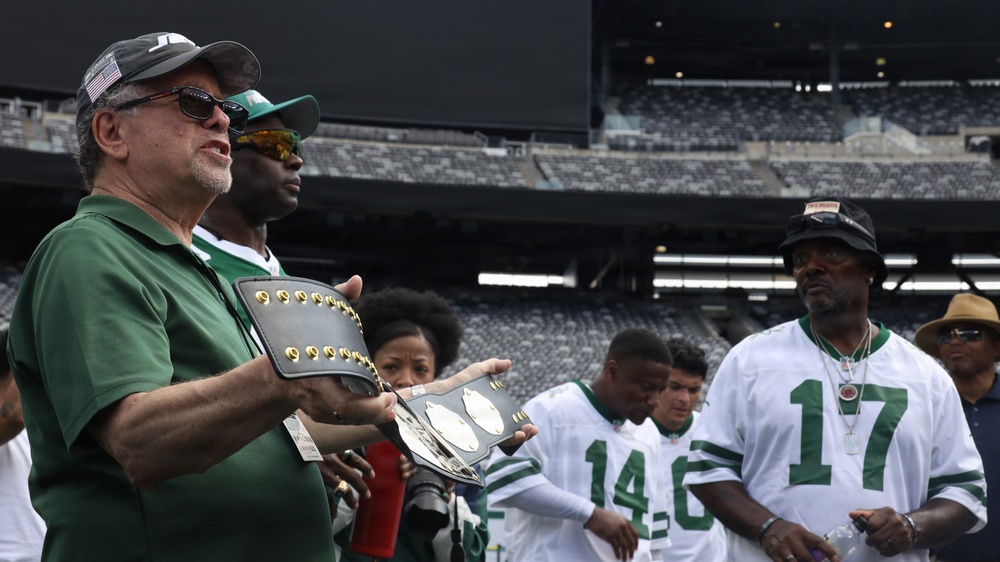 A group of people are gathered on a football field, likely at MetLife Stadium based on the visible seating and scoreboard. A man in a green polo shirt is holding what appears to be a championship belt or trophy plaque. Several individuals are wearing white and green jerseys consistent with New York Jets colors.