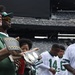 A group of people are gathered on a football field, likely at MetLife Stadium based on the visible seating and scoreboard. A man in a green polo shirt is holding what appears to be a championship belt or trophy plaque. Several individuals are wearing white and green jerseys consistent with New York Jets colors.