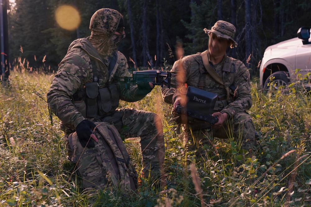 Two individuals in military camouflage uniforms are kneeling in a grassy field at the edge of a forested area during low-light conditions. One person is handling what appears to be a weapon or equipment, while the other holds a device labeled 'Parrot', which is a brand associated with military-grade drone systems. A civilian pickup truck is partially visible in the background.