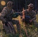 Two individuals in military camouflage uniforms are kneeling in a grassy field at the edge of a forested area during low-light conditions. One person is handling what appears to be a weapon or equipment, while the other holds a device labeled 'Parrot', which is a brand associated with military-grade drone systems. A civilian pickup truck is partially visible in the background.