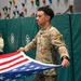 A U.S. Air Force service member in OCP (Operational Camouflage Pattern) uniform is handling/folding an American flag inside what appears to be a gymnasium or recreational facility. A young girl in a light-colored hoodie stands in the background observing. The setting includes a rock climbing wall with colorful holds and green padded safety mats.