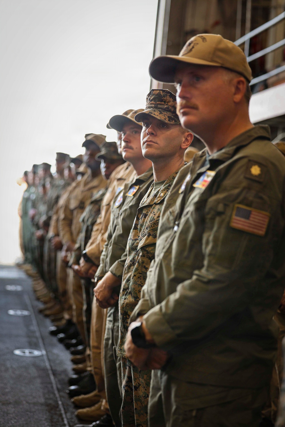 A formation of U.S. military personnel standing at attention in a line, appearing to be on the deck of a naval vessel. The group includes both U.S. Marines in MARPAT camouflage uniforms and personnel in olive drab flight suits. They appear to be participating in a formal ceremony or official event.
