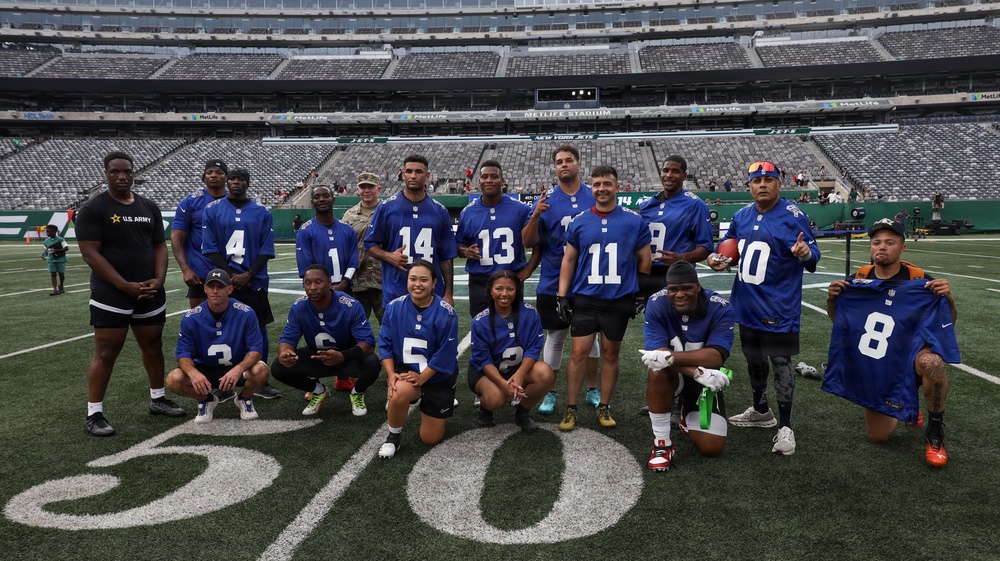 A group of approximately 18 people pose for a team photo on the field at MetLife Stadium, home of the New York Jets and New York Giants. Most participants are wearing blue New York Giants NFL jerseys with various numbers. The group includes military personnel, as evidenced by a U.S. Army shirt and military camouflage uniform visible among the group.
