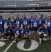 A group of approximately 18 people pose for a team photo on the field at MetLife Stadium, home of the New York Jets and New York Giants. Most participants are wearing blue New York Giants NFL jerseys with various numbers. The group includes military personnel, as evidenced by a U.S. Army shirt and military camouflage uniform visible among the group.