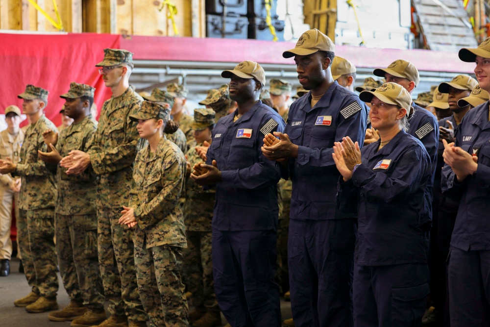 A group of U.S. military personnel in two distinct uniforms — Marine Corps MARPAT camouflage and Navy blue coveralls — are standing and applauding at what appears to be an indoor military ceremony or event. The setting appears to be inside a ship's hangar bay or large military facility, indicated by the industrial background with red curtains or barriers. Personnel are wearing tan/khaki caps and some display patches including what appears to be a Texas state flag patch.