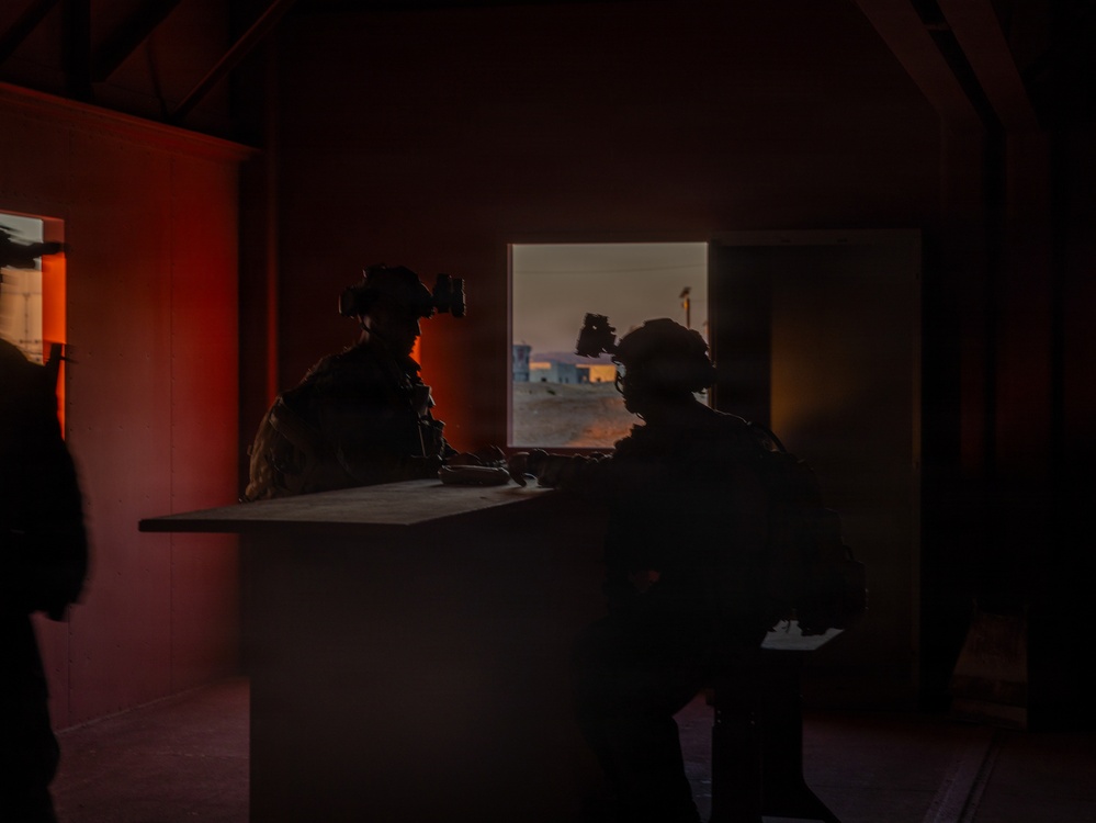 Two silhouetted military personnel wearing helmets equipped with night vision devices are positioned inside a darkened structure, observing through an open window or doorway. The exterior visible through the opening shows a arid/desert environment at dusk or dawn with warm orange light. The interior is dimly lit with a reddish ambient glow from the left side.