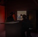Two silhouetted military personnel wearing helmets equipped with night vision devices are positioned inside a darkened structure, observing through an open window or doorway. The exterior visible through the opening shows a arid/desert environment at dusk or dawn with warm orange light. The interior is dimly lit with a reddish ambient glow from the left side.