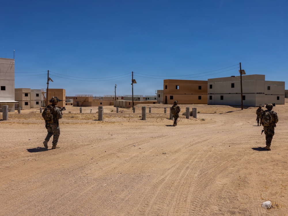 The image shows military personnel in full combat gear patrolling through what appears to be a mock urban training environment in a desert setting. The area consists of unoccupied concrete and tan-colored multi-story buildings arranged along sandy dirt roads, consistent with a military training facility. The scene suggests an urban warfare or patrol training exercise taking place under clear, sunny desert conditions.
