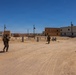 The image shows military personnel in full combat gear patrolling through what appears to be a mock urban training environment in a desert setting. The area consists of unoccupied concrete and tan-colored multi-story buildings arranged along sandy dirt roads, consistent with a military training facility. The scene suggests an urban warfare or patrol training exercise taking place under clear, sunny desert conditions.