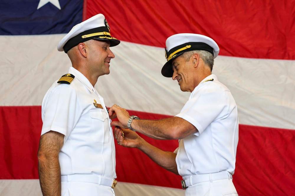 Two U.S. Navy officers in white dress uniforms are participating in a formal military decoration or pinning ceremony. A senior officer is pinning a medal or insignia onto the chest of a junior officer, both smiling, with a large American flag displayed prominently in the background. The setting appears to be an official military ceremony held indoors.