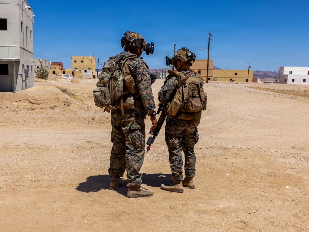 Two military personnel in full combat gear are standing in a desert training environment, viewed from behind. They are equipped with digital camouflage uniforms, tactical vests, helmets with mounted optics, and backpacks. The setting appears to be a military urban warfare training facility with constructed buildings in an arid desert landscape.