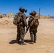Two military personnel in full combat gear are standing in a desert training environment, viewed from behind. They are equipped with digital camouflage uniforms, tactical vests, helmets with mounted optics, and backpacks. The setting appears to be a military urban warfare training facility with constructed buildings in an arid desert landscape.